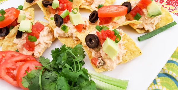 Chicken Tinga Tostaditas-shredded chicken with chipotle mayo, onions, tomatoes, scallions, cilantro, black olives, avocado on top of tortilla chips and served on a white platter.