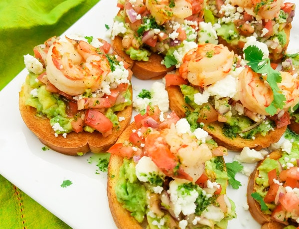 This Mexican Bruschetta Recipe is made with a fresh guacamole, tomatoes, red onions, jalapeños, garlic, cilantro, lime juice, topped with queso fresco and savory cooked shrimp.  (Shrimp optional)  Excellent snack or starter to any meal. Served on top of a white platter. 