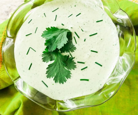 A poblano cream sauce made with roasted poblano peppers, sour cream, mayonnaise, garlic, lime juice, cilantro, salt and pepper inside a glass dipping cup and garnished with cilantro leaves and chives.