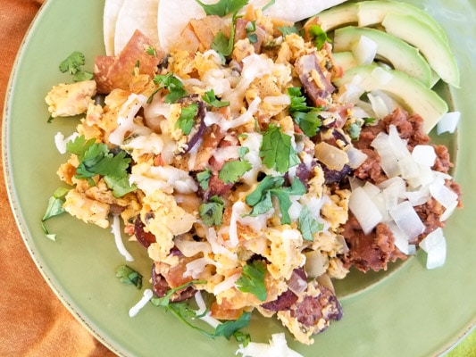 Authentic Migas Recipe with crispy corn tortilla strips, scrambled eggs with kielbasa sausage, onions, tomatoes, poblano pepper, jalapeno, garlic, cilantro, monterey jack cheese and refried beans, avocado slices on a green plate.