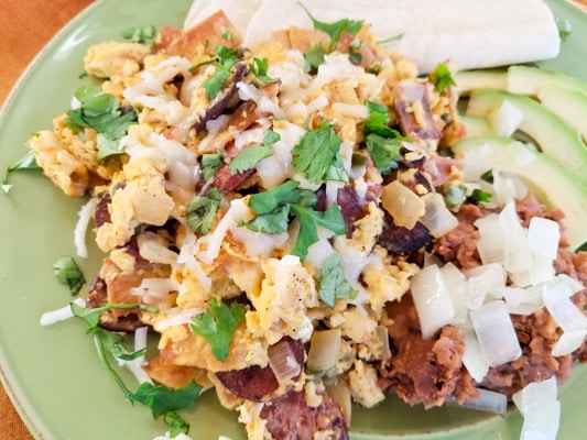 Authentic Migas Recipe with crispy corn tortilla strips, scrambled eggs with kielbasa sausage, onions, tomatoes, poblano pepper, jalapeno, garlic, cilantro, monterey jack cheese and refried beans, avocado slices on a green plate.