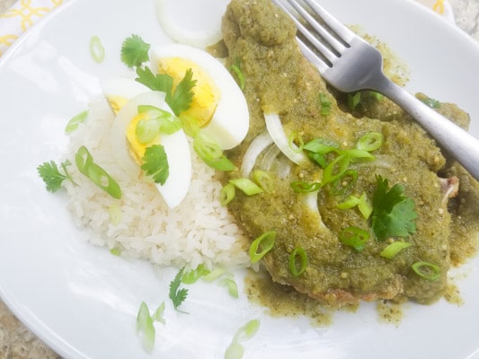 Chuletas de Puerco or Chuletas de Puerco en Salsa Verde (Pork Chops in Salsa Verde served with white rice, thin slices of onions and a quartered hard boiled egg sitting on top and served on a white plate.