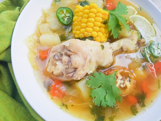 Caldo de Pollo (Mexican Chicken Soup) with carrots, onion, green cabbage, corn cob pieces and jalapenos served in a white bowl.