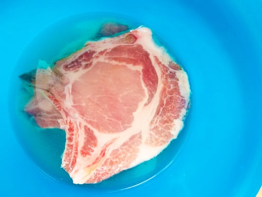 Pork chops sitting in a brine solution in a large blue bowl.
