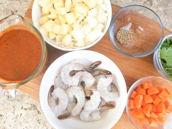 Ingredients ready for the soup. Tomato guajillo puree, cubed potatoes, chopped carrots, deshelled shrimps and spices in separate bowls on top of a wooden cutting board.