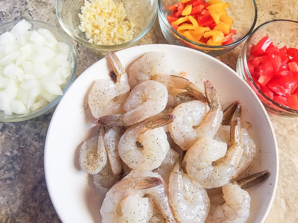 Shrimps, chopped onions, peppers, tomatoes and garlic in individual bowls on a wooden cutting board.
