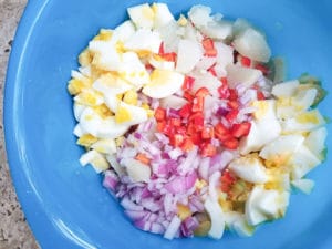 Red onions, red peppers, boiled eggs chopped with cooked potatoes in a blue boil to make Puerto Rican potato salad or ensalada de papa.