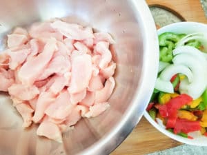 Raw chicken in a steel bowl with a bowl of mixed peppers ans onions beside it.