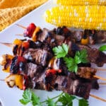 Alambres de Res (Beef Kabobs) served on a white platter with a side of corn on the cob.