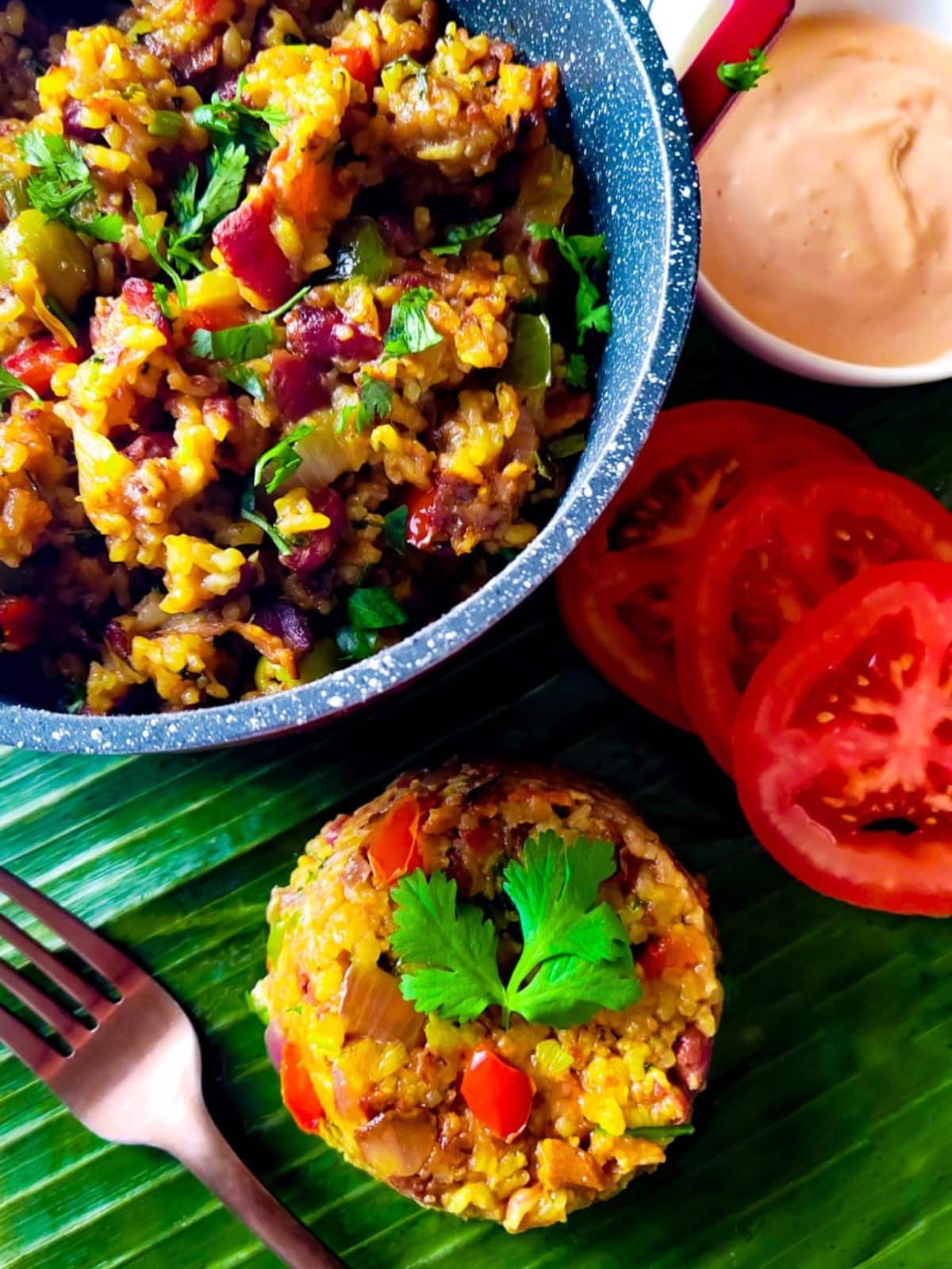 Arroz Mamposteao served on top of a plantain leaf with a side of tomato slices, lime wedges and a pot of rice beside it all.