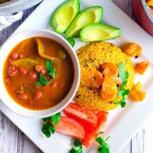 Arroz con Camarones (Puerto Rican Rice and Shrimps)served on a white plate with a side of habichuelas guisadas (puerto Rican Stewed Beans), avocado slices, and tomatoes slices.