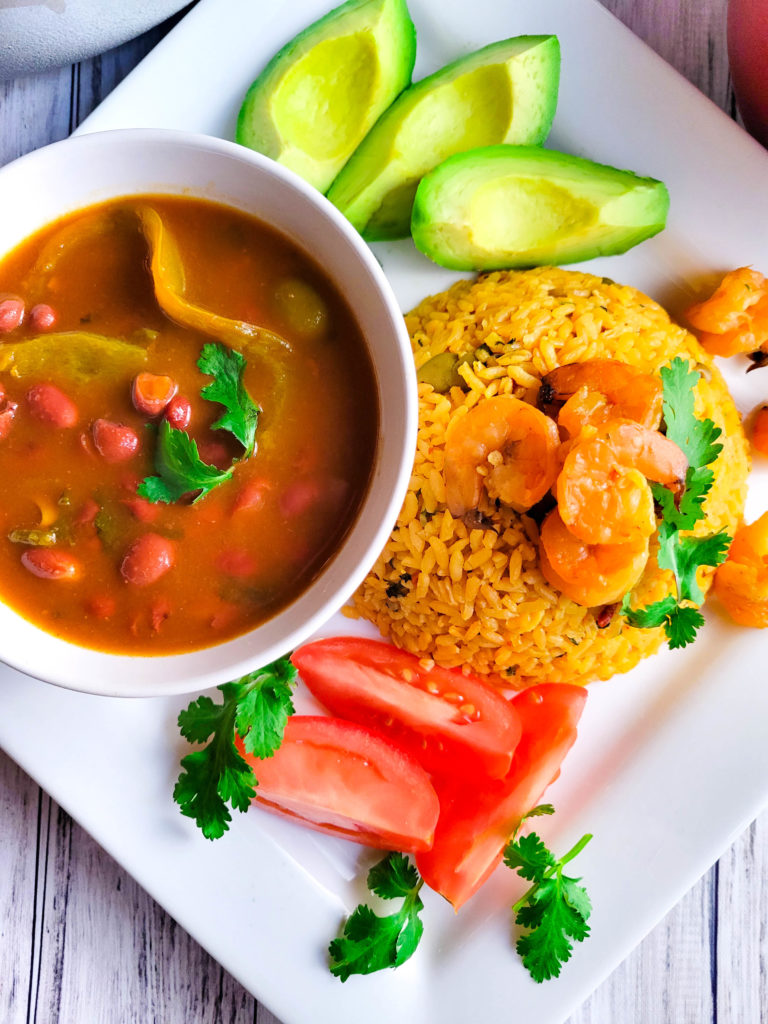 Arroz con Camarones (Puerto Rican Rice and Shrimps)served on a white plate with a side of habichuelas guisadas (Puerto Rican Stewed Beans), avocado slices, and tomatoes slices.