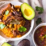 Puerto Rican arroz con costillas served on a white bowl with a side of habichuelas guisadas and avocado.