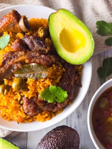 Puerto Rican arroz con costillas served on a white bowl with a side of habichuelas guisadas and avocado.