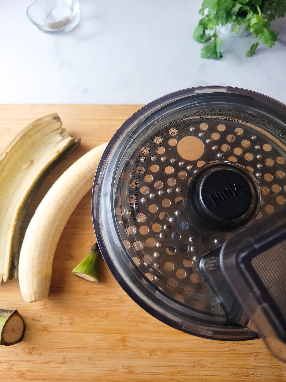 A plantain peeled on a wooden cutting board with a food processor beside it to blend the plantain into a puree.