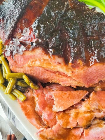 A fully cooked Bourbon Maple Glazed Ham served on a large white platter with a side of green beans.