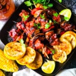 Brochetas de Pollo (Chicken Kabobs) cooked in a cast iron grilled skillet with slices of yellow squash on skillet, corn on the cobs on the side on a plate and a mason jar with guava barbecue sauce beside skillet.
