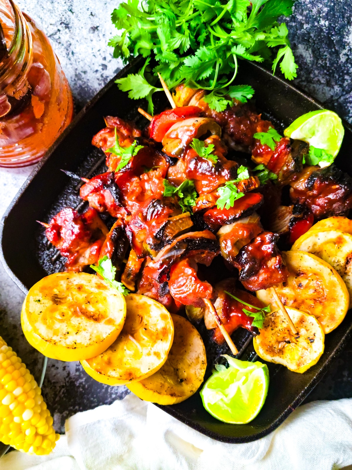 Brochetas de Pollo (Chicken Kabobs) cooked in a cast iron grilled skillet with slices of yellow squash on skillet, corn on the cobs on the side on a plate and a mason jar with guava barbecue sauce beside skillet.