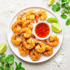 Camarones coco (coconut shrimp) served on a white round plate with chili duck sauce and lime wedges.