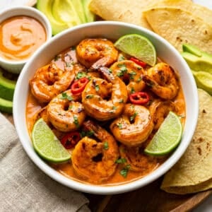 Camarones al chipotle served in a white bowl with lime wedges and served on a round wooden cutting board with a side of avocado slices and corn tortillas.