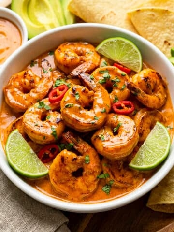Camarones al chipotle served in a white bowl with lime wedges and served on a round wooden cutting board with a side of avocado slices and corn tortillas.