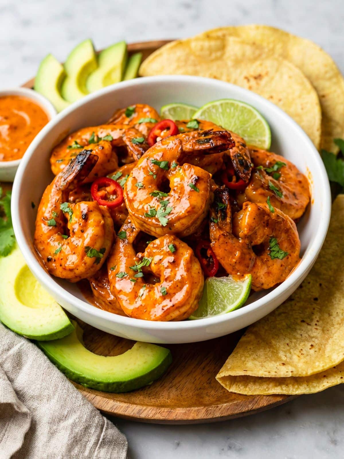 Camarones al chipotle served in a white bowl with lime wedges and served on a round wooden cutting board with a side of avocado slices and corn tortillas.