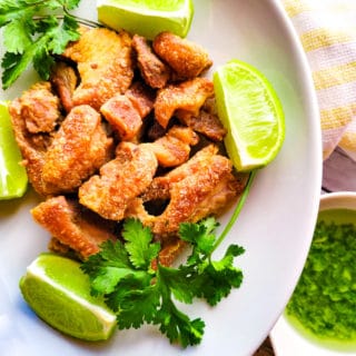 Chicharrones de Puerco (Pork Cracklings) served on a white plate with cilantro garlic aioli and lime wedges.