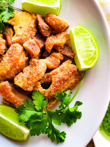 Chicharrones de Puerco (Pork Cracklings) served on a white plate with cilantro garlic aioli and lime wedges.