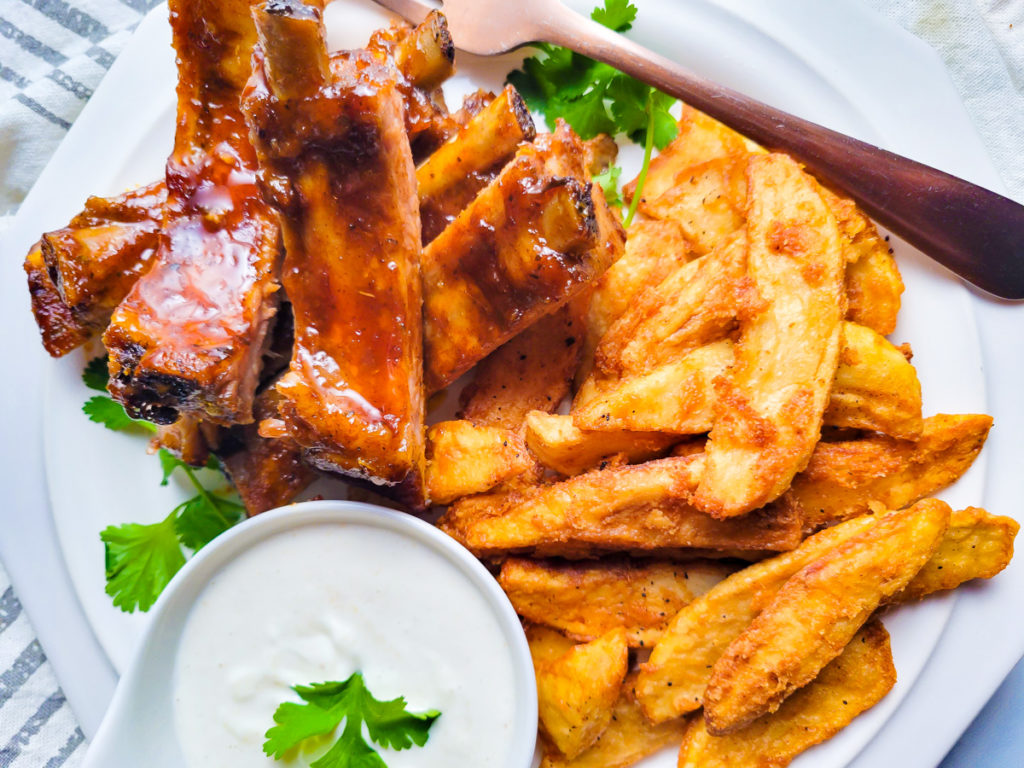 Costillas en Barbecue served with seasoned fries on a white plate with a side of horseradish sauce.