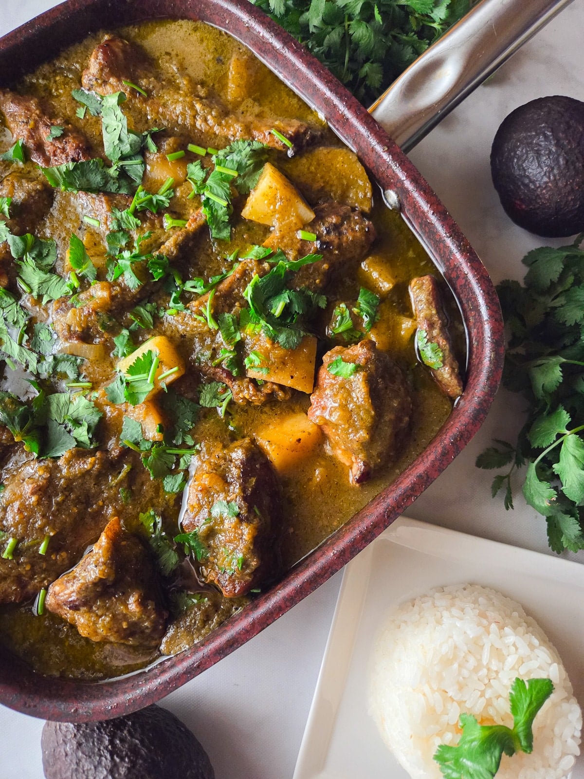 Costillas en salsa verde cooked in a large saucepan with a side of white rice and avocados.