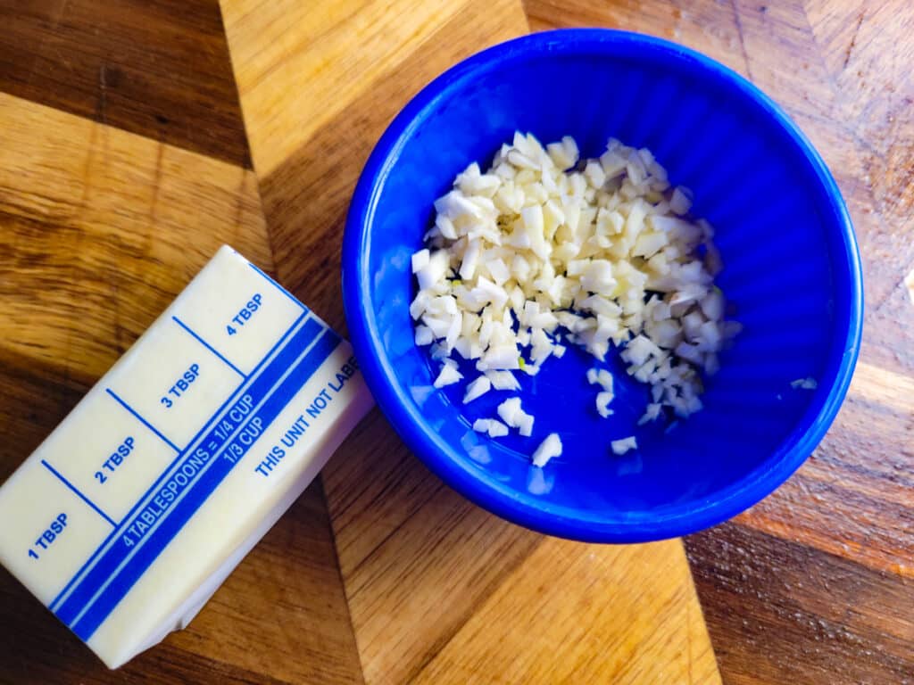 Chopped garlic in a small condiment bowl and half a stick of butter on a wooden cutting board.