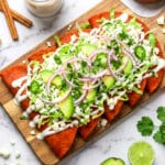 Enchiladas Potosinas served on a wooden cutting board, topped with shredded lettuce, avocado, red onions, and jalapenos. Beside it, a glass of Mexican horchata and avocado.