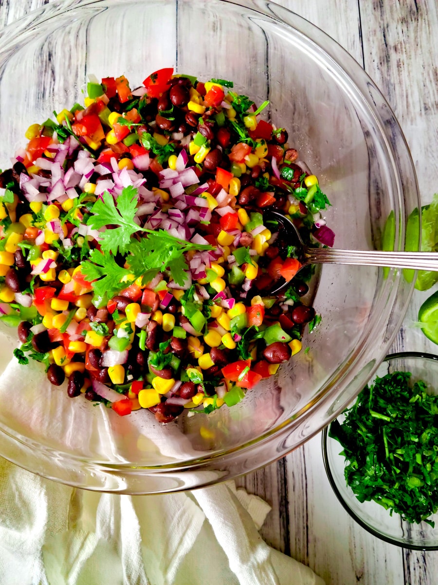 Ensalada de Frijoles Negros (Black Bean Salad) in a mixing bowl with chopped cilantro and lime wedges on the side.