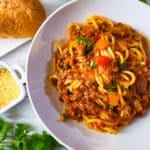 Espagueti con carne molida (Puerto Rican Spaghetti) served in a white bowl with a side of bread, tossed salad and grated parmesan cheese.