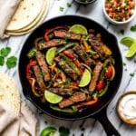 Fajitas de res cooked in a cast iron skillet surrounded by toppings and flour tortillas.