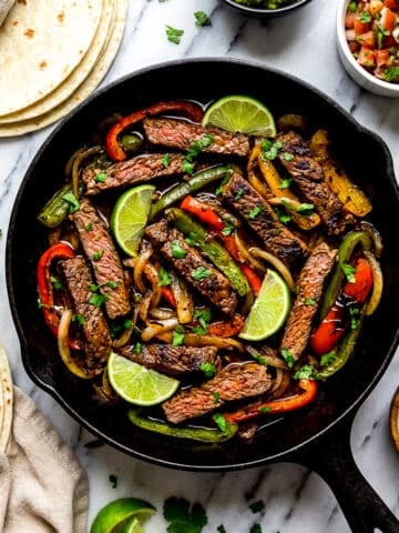 Fajitas de res cooked in a cast iron skillet surrounded by toppings and flour tortillas.