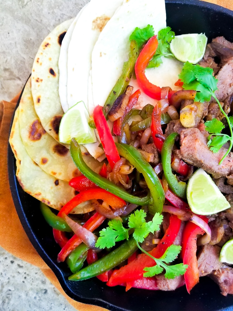 Fajitas de Res (Beef Fajitas) served in a cast iron skillet with green, red peppers, onions, burrito and corn wraps, topped with cilantro sprigs and lime wedges.