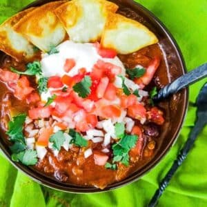 An amazing ground venison chili served in a brown bowl. This ground venison chili is made with venison and a blend of onions, peppers, jalapenos,garlic, black beans, kidney beans in a savory tomato sauce and topped with sour cream, chopped onions, chopped tomatoes, cilantro and homemade tortilla chips.