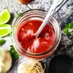Guava BBQ Sauce (Salsa de Guayaba) in a mason jar with whole heads of garlic beside and lime wedges.