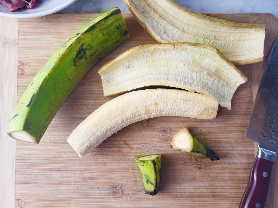 Green plantains on a wooden cutting board, ends cut and peeled.
