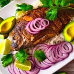 Mojarra frita laying on top of a bed of white rice surrounding by slices of red onions and avocado slices.