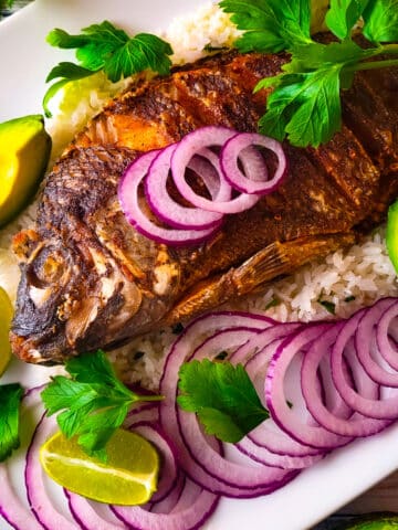 Mojarra frita laying on top of a bed of white rice surrounding by slices of red onions and avocado slices.