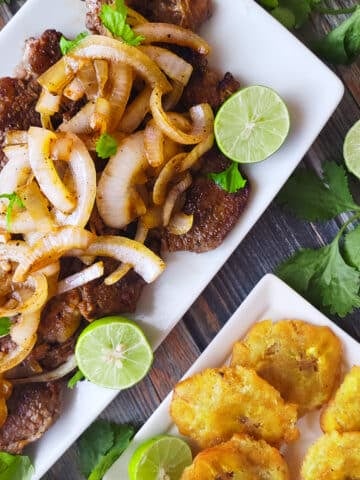 Palomilla steak served on a white platter topped with onions and a side of tostones (fried plantains).