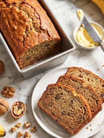Pan de platano (pan de banana) in a loaf pan with a few banana bread slices on a white plate. Ripe bananas, butter and walnuts around bread for decor.