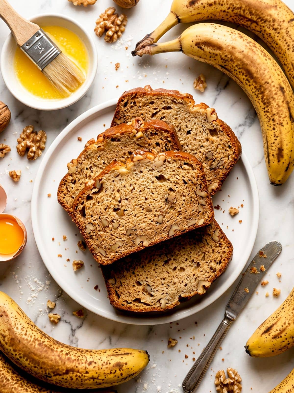Pan de platano (pan de banana) slices on a white plate. Ripe bananas, butter and walnuts around bread for decor.