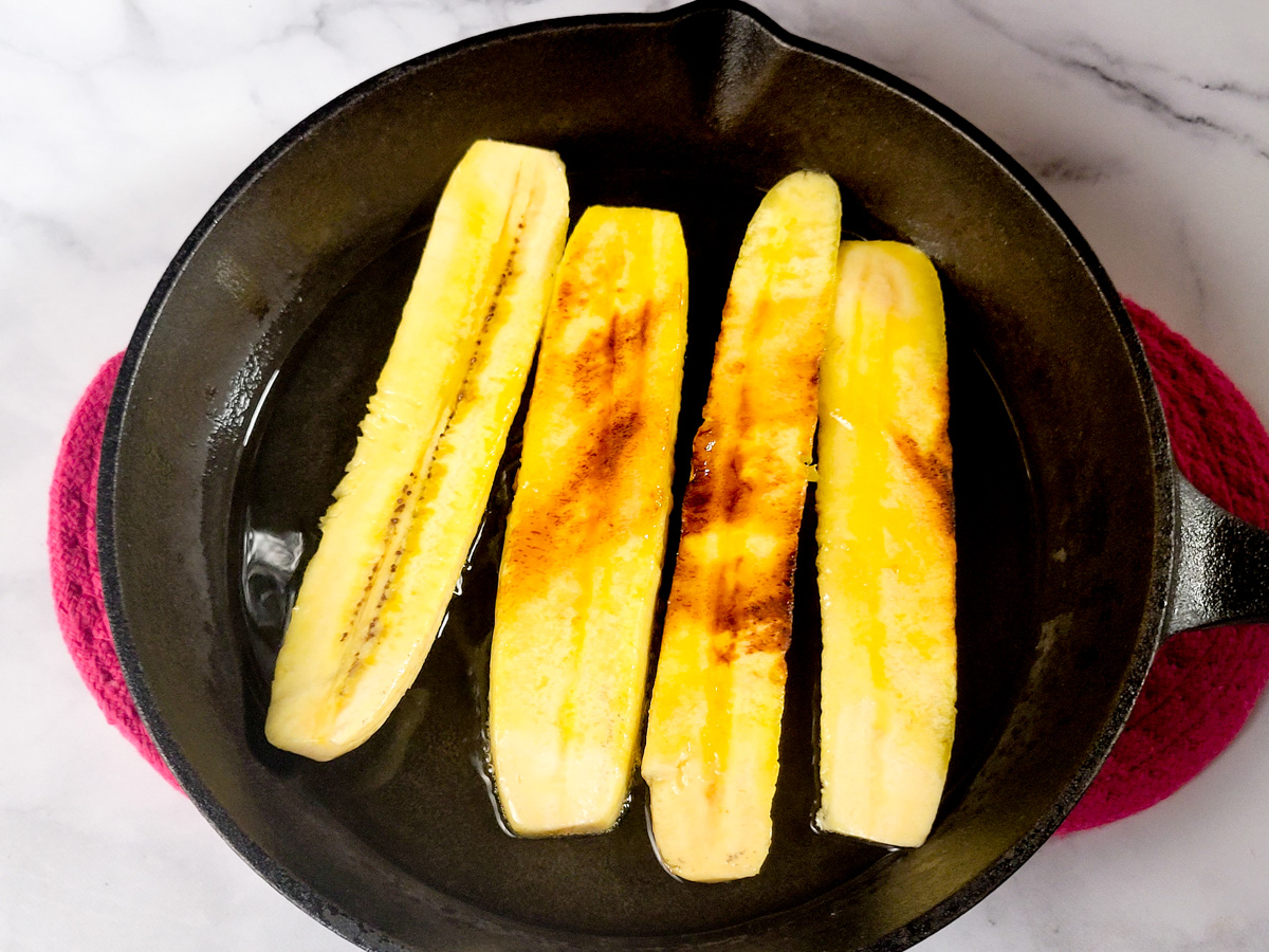 Vertically sliced plantains frying in a cast iron skillet.