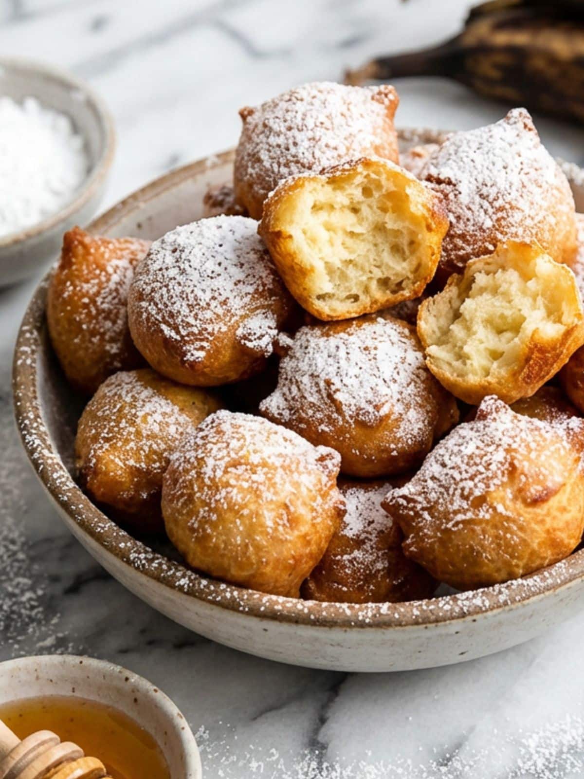 Plantain Puff Puff served in a white bowl and topped with confectioners sugar. A small bowl of ground cinnamon and honey sit beside the bowl.