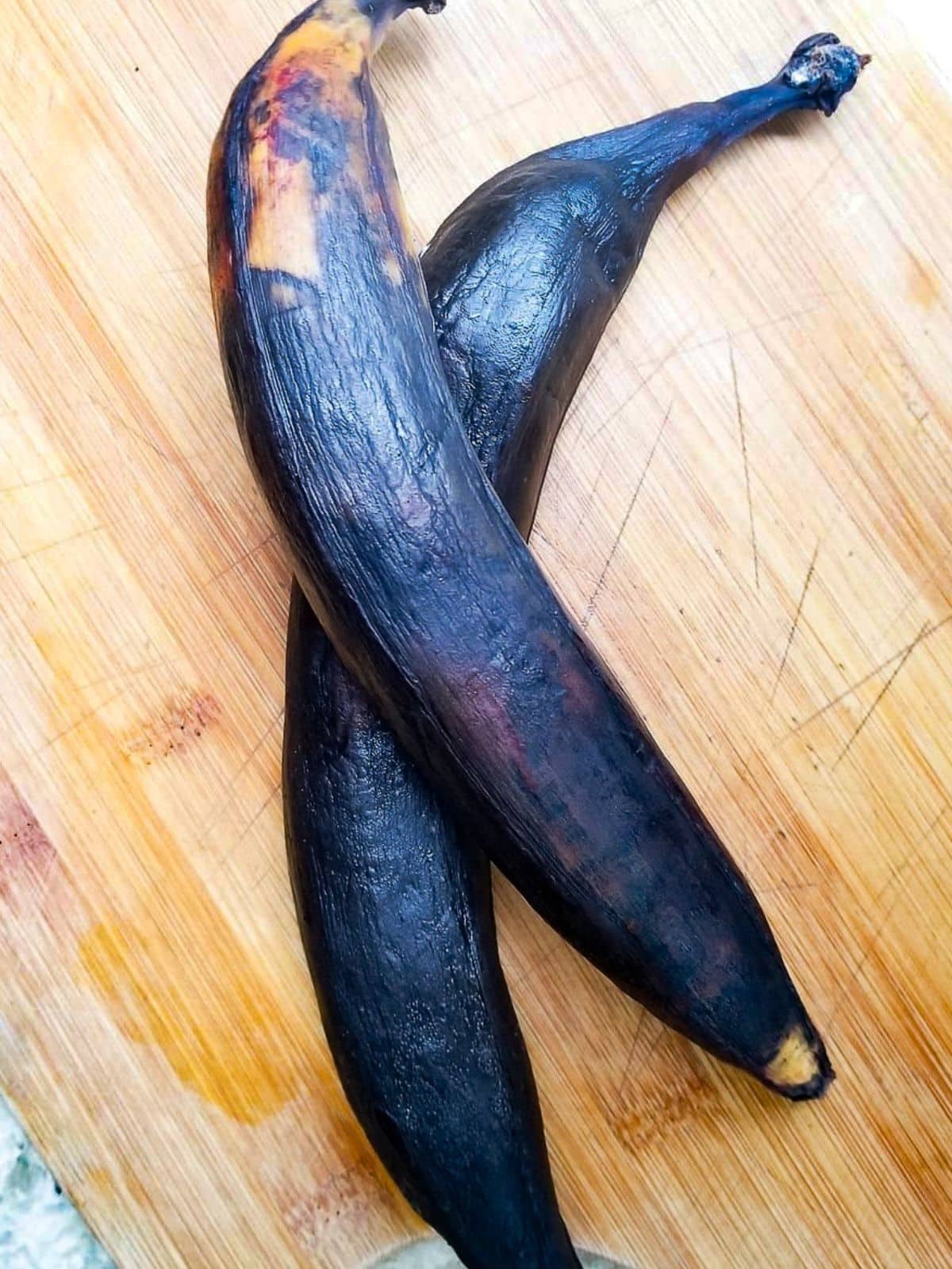 Overripe black skinned plantains sitting on top of a wooden cutting board.