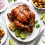 Pollo Rostizado (Rotisserie Chicken) served on a white platter with lime wedges, whole roasted garlic and cilantro garnish.