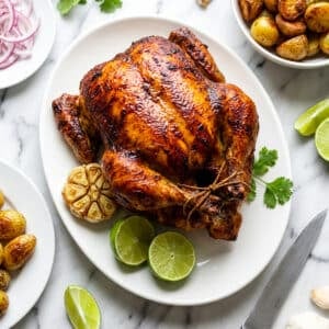 Pollo Rostizado (Rotisserie Chicken) served on a white platter with lime wedges, whole roasted garlic and cilantro garnish.